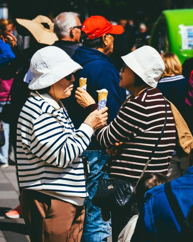 Two adults enjoying ice cream cones outdoors in a bustling crowd during a sunny day.